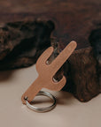 A cactus-shaped metal keychain tool with a bottle opener cutout and a silver key ring, photographed on a beige surface with dark wood in the background.