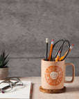 Decorative mug with sun design filled with office supplies on a wooden surface with a plant and glasses in the background.