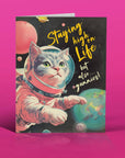 Card with a cat in an astronaut suit and space-themed text on a pink background
