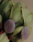 A pair of gold drop earrings with oval lavender-purple stone pendants, displayed on the green petals of an artichoke against a soft beige background.