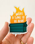 Sticker with a dumpster fire design and text held by a hand on a light background