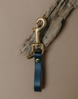 A dark blue leather key ring strap with a gold-tone metal swivel clasp and key ring, displayed against a neutral beige background with a piece of driftwood.