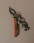 A brown leather key fob or keychain with a brass key ring, hanging from a piece of weathered driftwood against a solid taupe background.