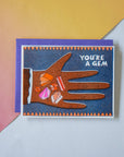 Greeting card with a hand holding gemstones and 'You're a Gem' text on a colorful background