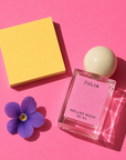 Julia perfume bottle beside a purple violet flower and a yellow block on a pink background