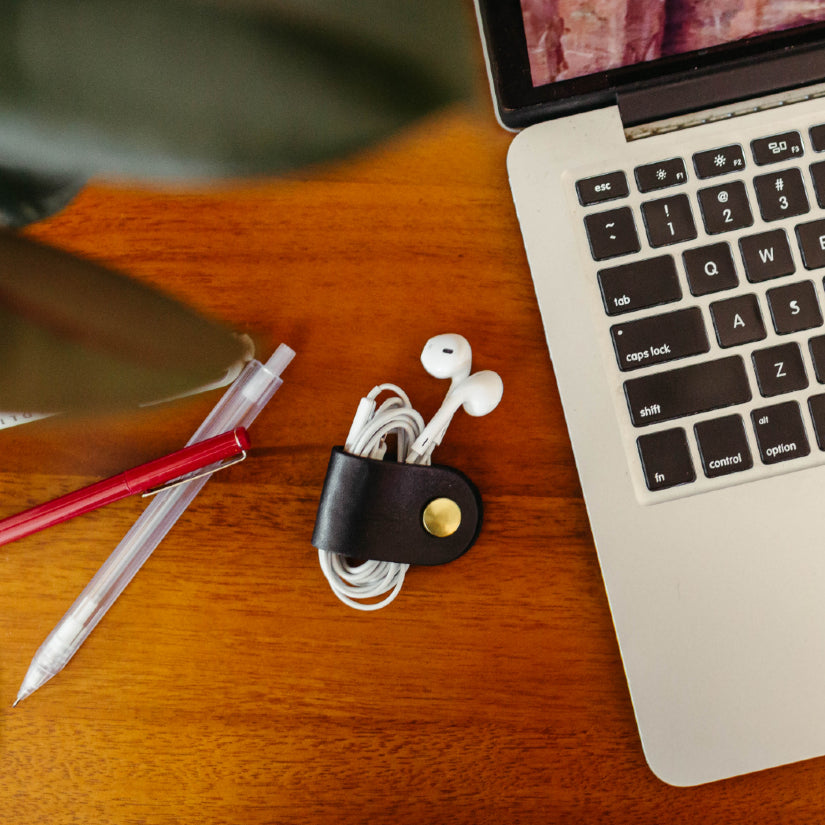 Black leather cord keeper holding earbuds on a wooden desk beside a laptop and pens