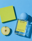 Betsey perfume bottle beside half a green apple and a chartreuse block on a blue background