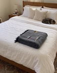 Folded charcoal wool thunderbird blanket on a white bed with a cat curled up near the pillows