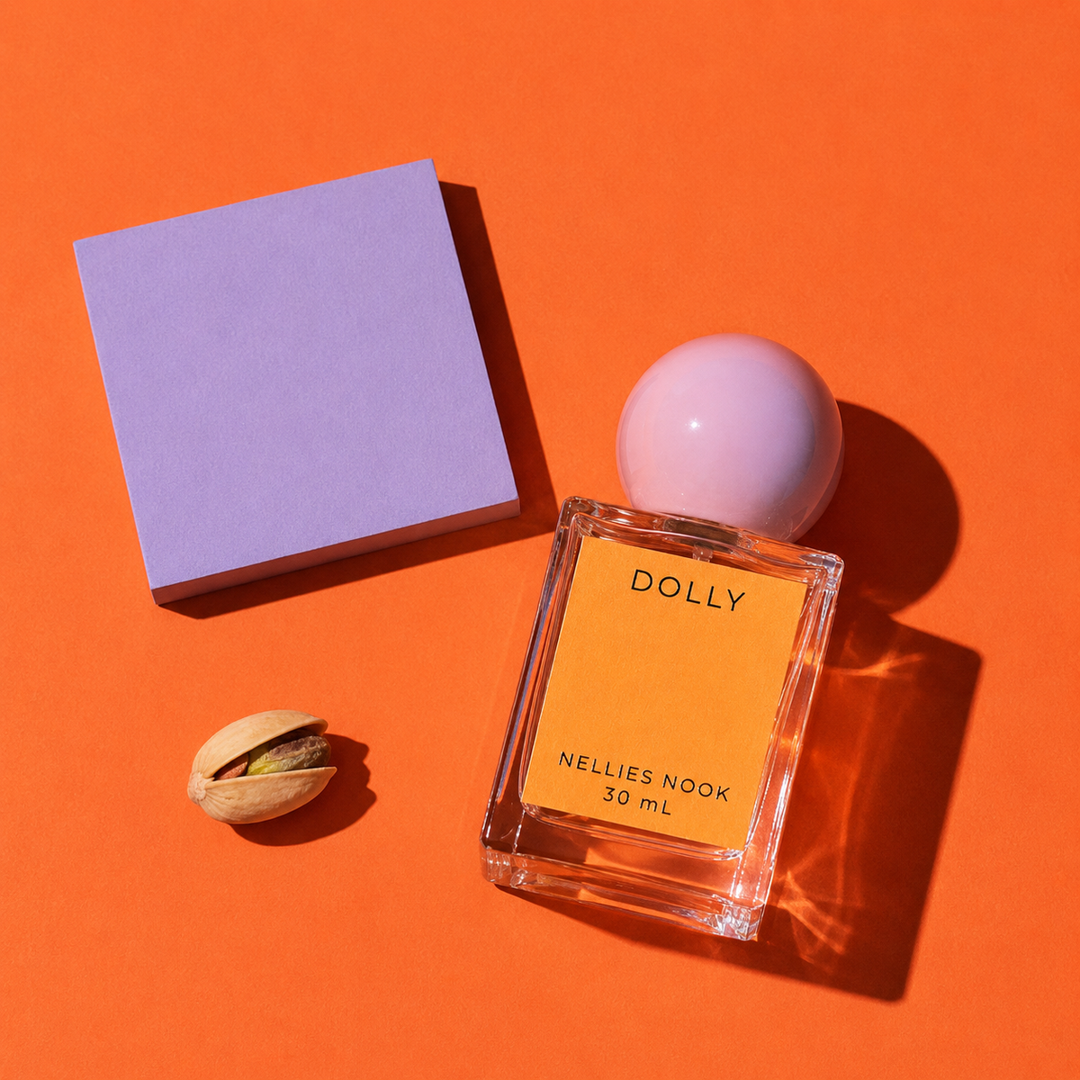 Dolly perfume bottle beside a pistachio and a lavender block on an orange background