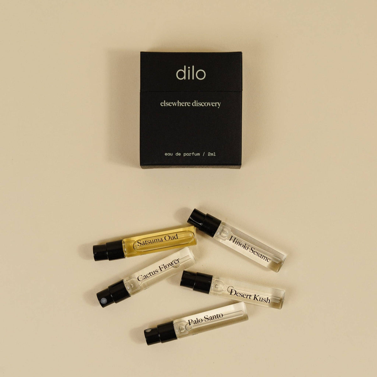 Five small perfume sample vials with black spray caps are arranged on a warm beige background below a black rectangular box labeled 'dilo elsewhere discovery eau de parfum / 5 ml'. The vials contain clear, yellow, and white liquids, with some labels partially visible including 'Sakura Oud', 'Citrus Flower', 'Desert Kush', and 'Pale Samso'.