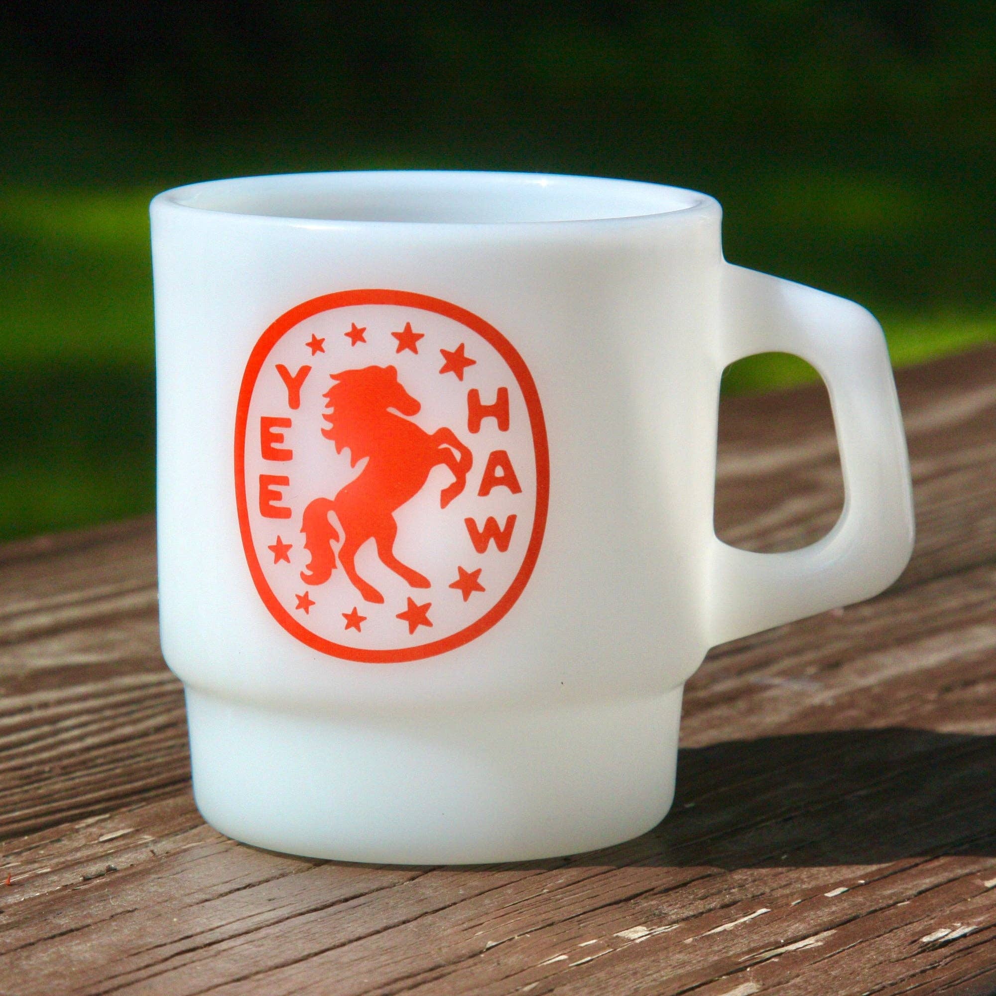 A white ceramic coffee mug with a red circular 'YEE HAW' horse logo, resting on a weathered wooden surface with a blurred green background.