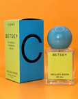 Green and blue Icons Betsey box beside a glass perfume bottle with a light blue sphere cap and green label, on an orange background