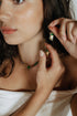 Model in a white top touching her ear, wearing a small crystal hoop and a beaded necklace