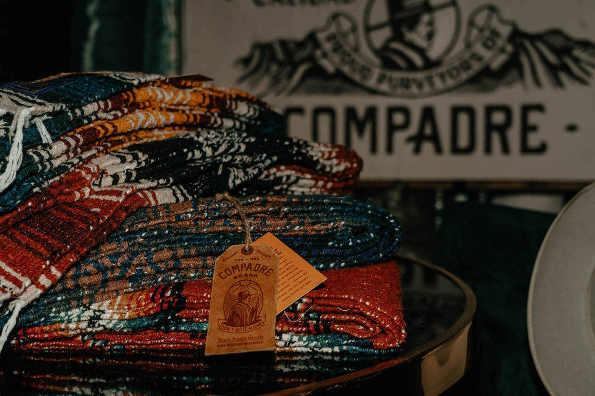 Stack of folded striped Compadre blankets with a hang tag, in front of a Compadre sign