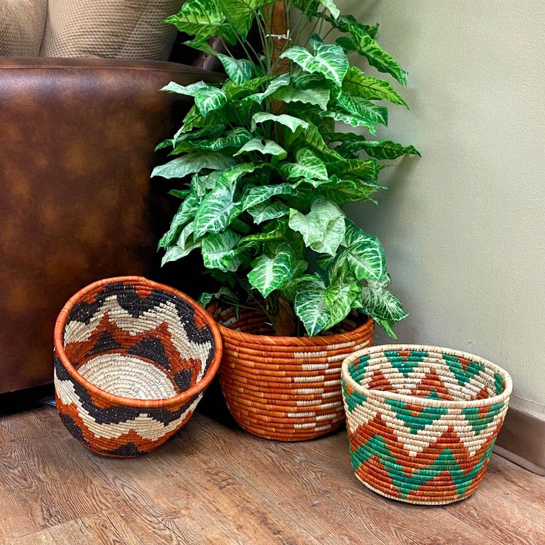 Three woven planter baskets in rust, black and green patterns beside a large potted plant and a leather chair