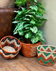 Three woven planter baskets in rust, black and green patterns beside a large potted plant and a leather chair