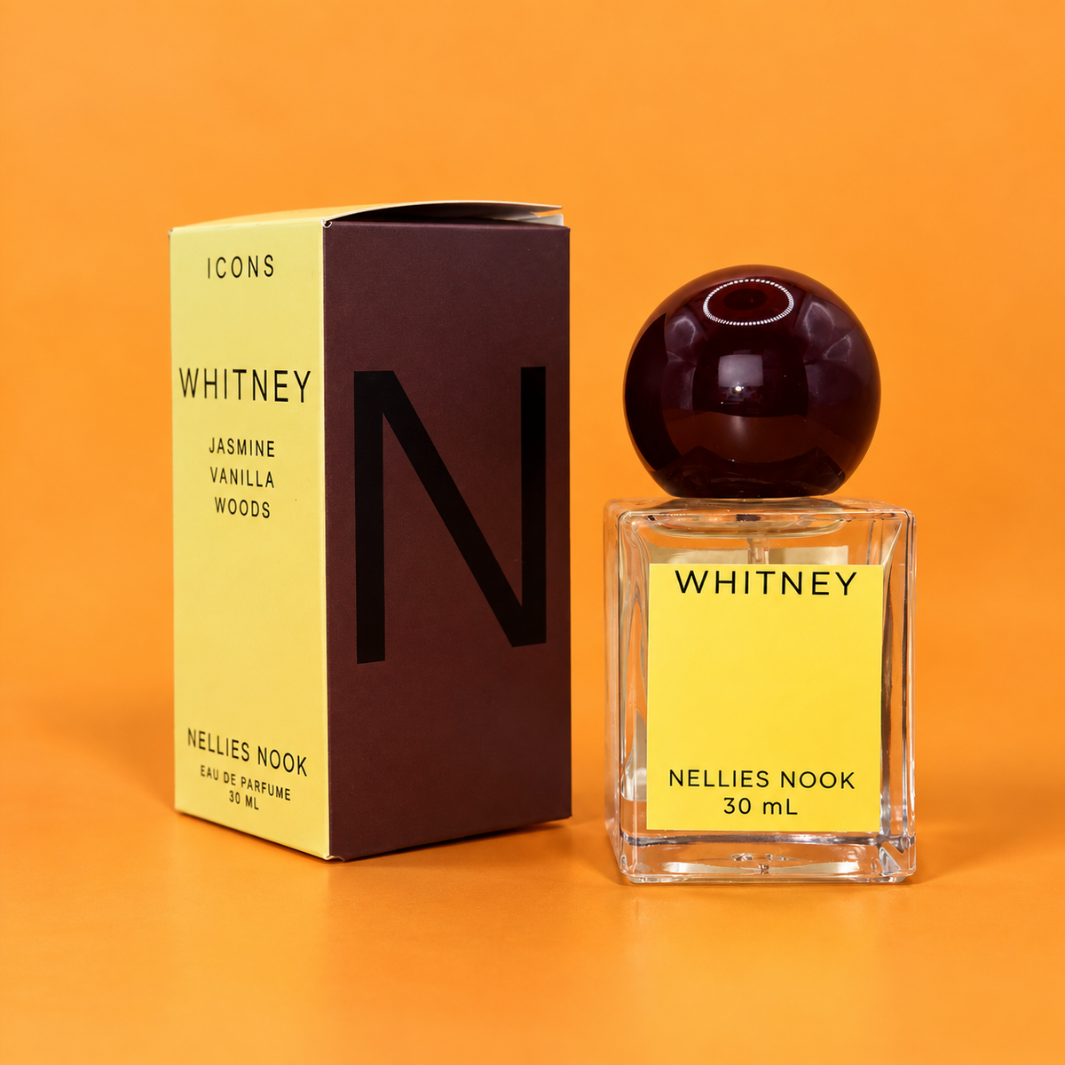 Yellow and brown Icons Whitney box beside a glass perfume bottle with a dark burgundy sphere cap and yellow label, on an orange background