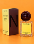 Yellow and brown Icons Whitney box beside a glass perfume bottle with a dark burgundy sphere cap and yellow label, on an orange background