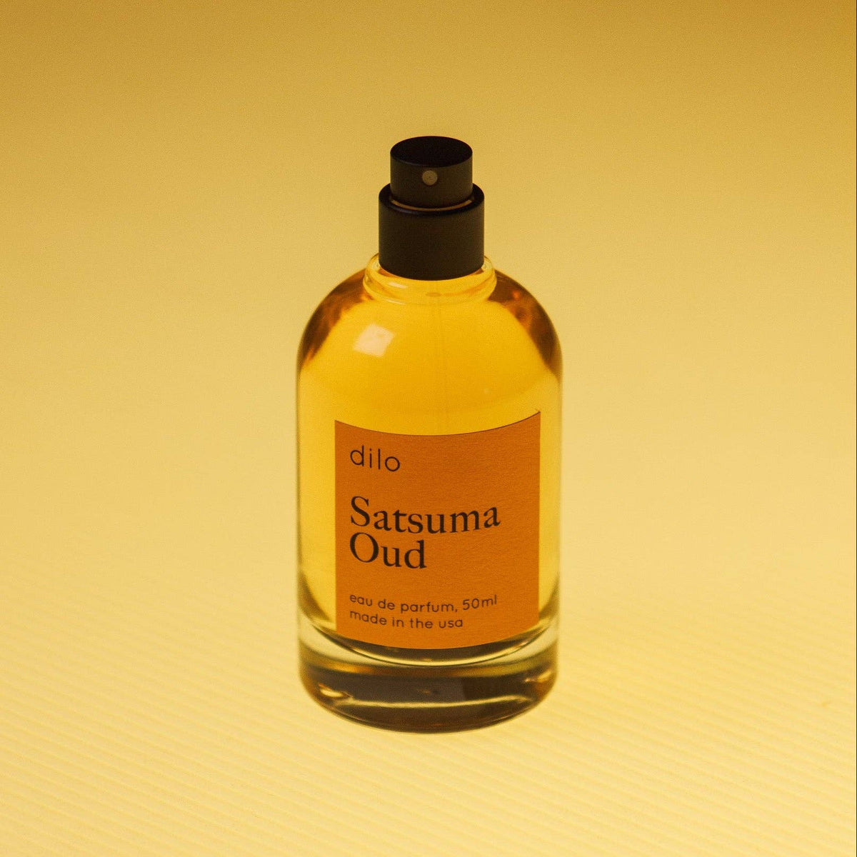 A 50ml bottle of 'dilo' Satsuma Oud Eau de Parfum with amber-colored liquid, a black spray nozzle, and an orange label, set against a warm yellow gradient background.