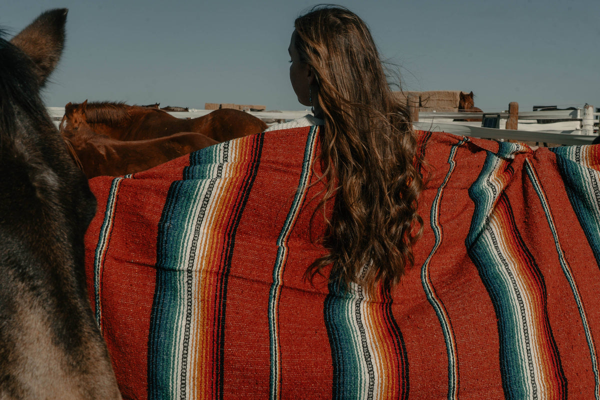 Person with long hair wrapped in a red and teal striped sarape beside a horse