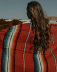 Person with long hair wrapped in a red and teal striped sarape beside a horse