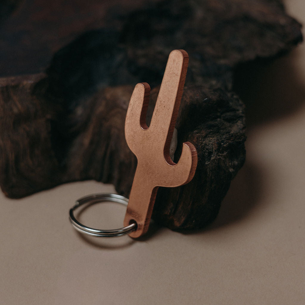 A cactus-shaped keychain made of copper-colored metal with a silver split key ring, resting against a dark textured wood prop on a beige background.
