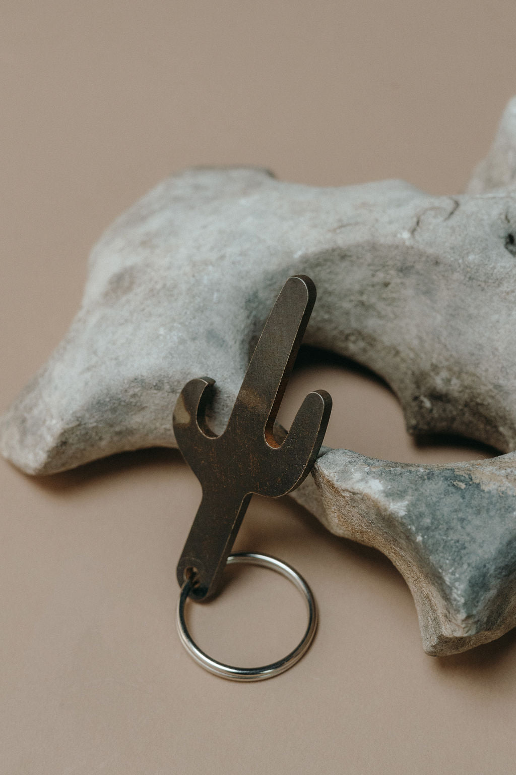 A cactus-shaped metal keychain tool with a silver split ring, resting against a light gray decorative bone on a warm beige background.