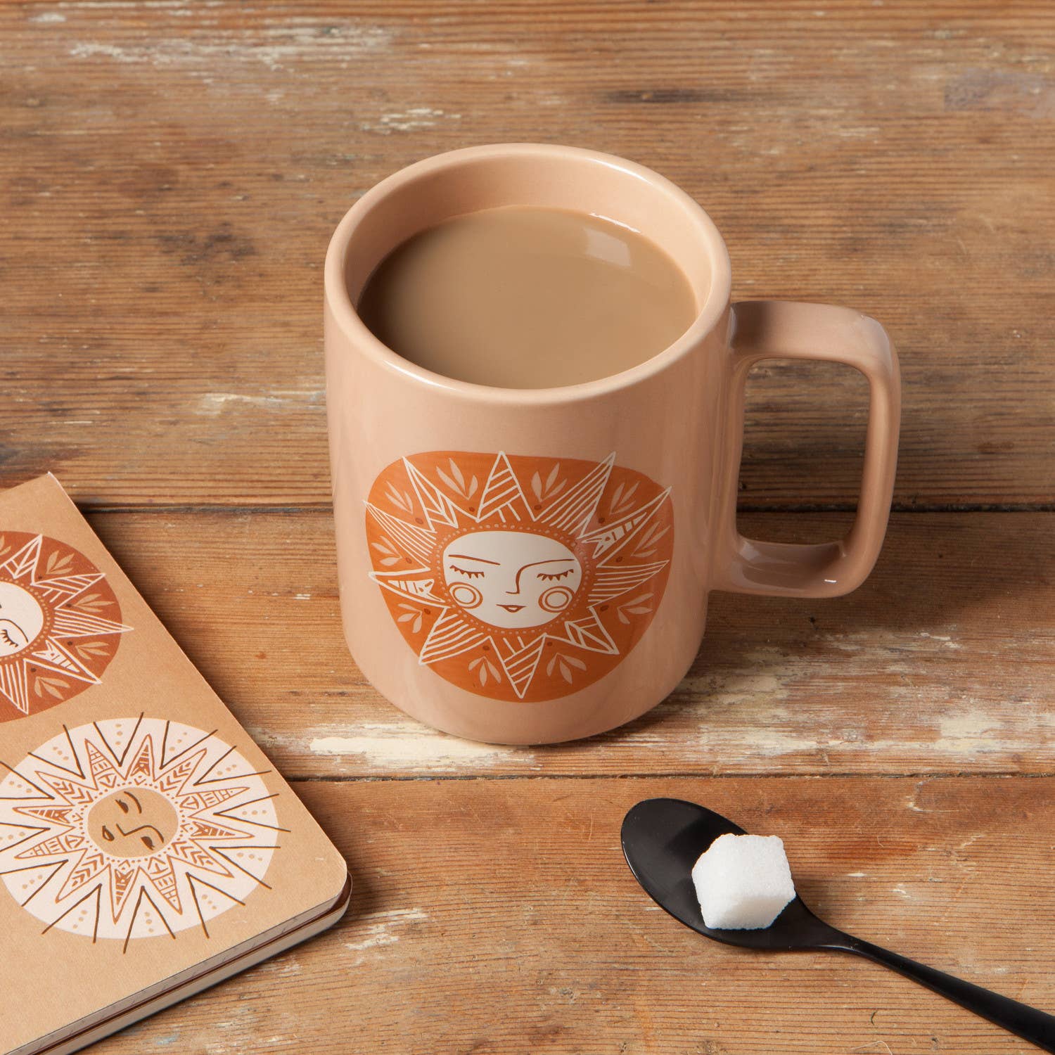 Ceramic mug with sun design on a wooden surface, next to a notebook and spoon with sugar.