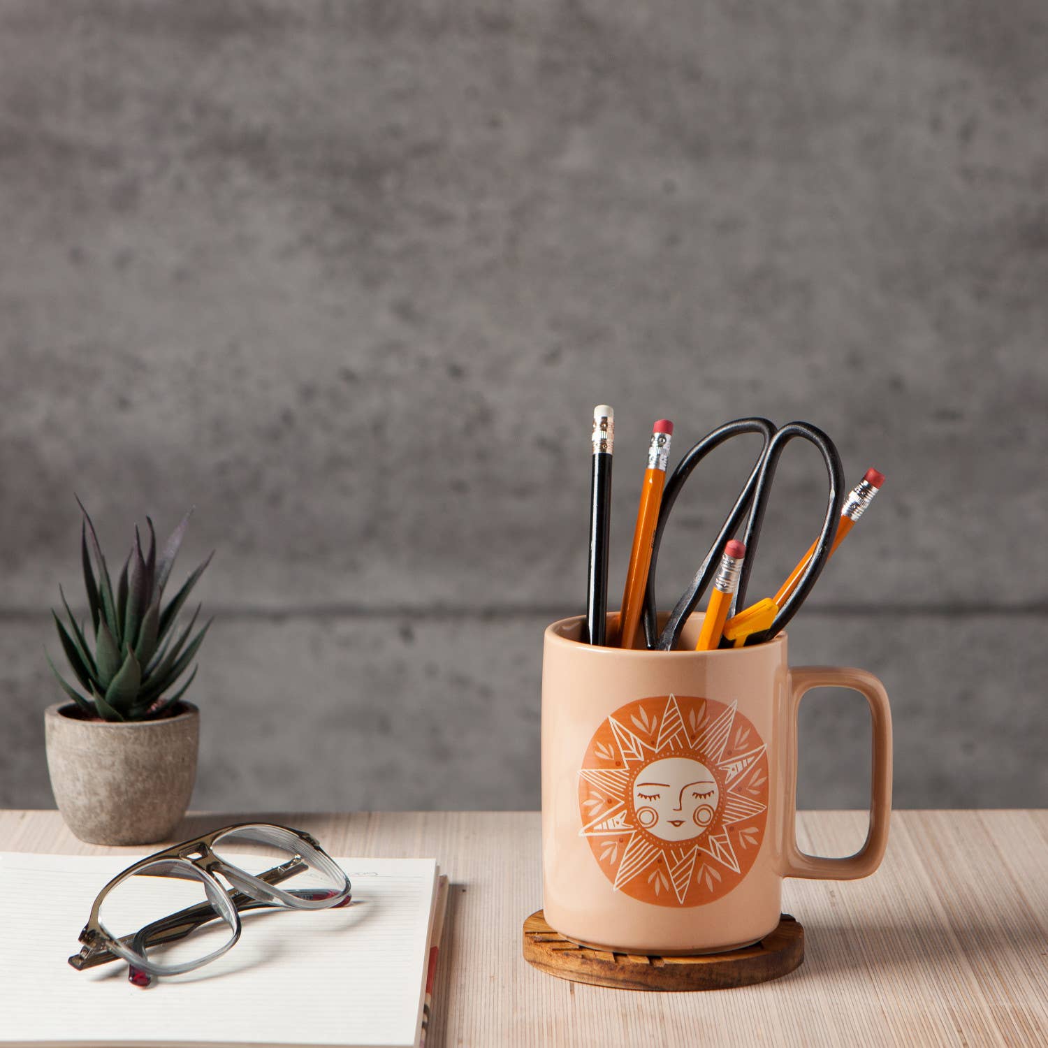 Decorative mug with sun design filled with office supplies on a wooden surface with a plant and glasses in the background.