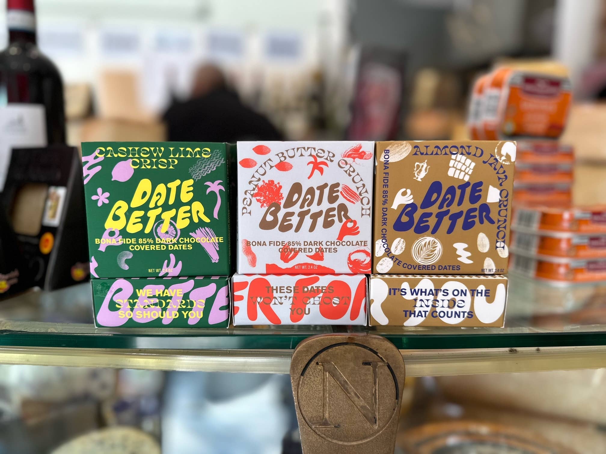 Three 'Date Better' chocolate packaging boxes on a glass shelf with blurred background