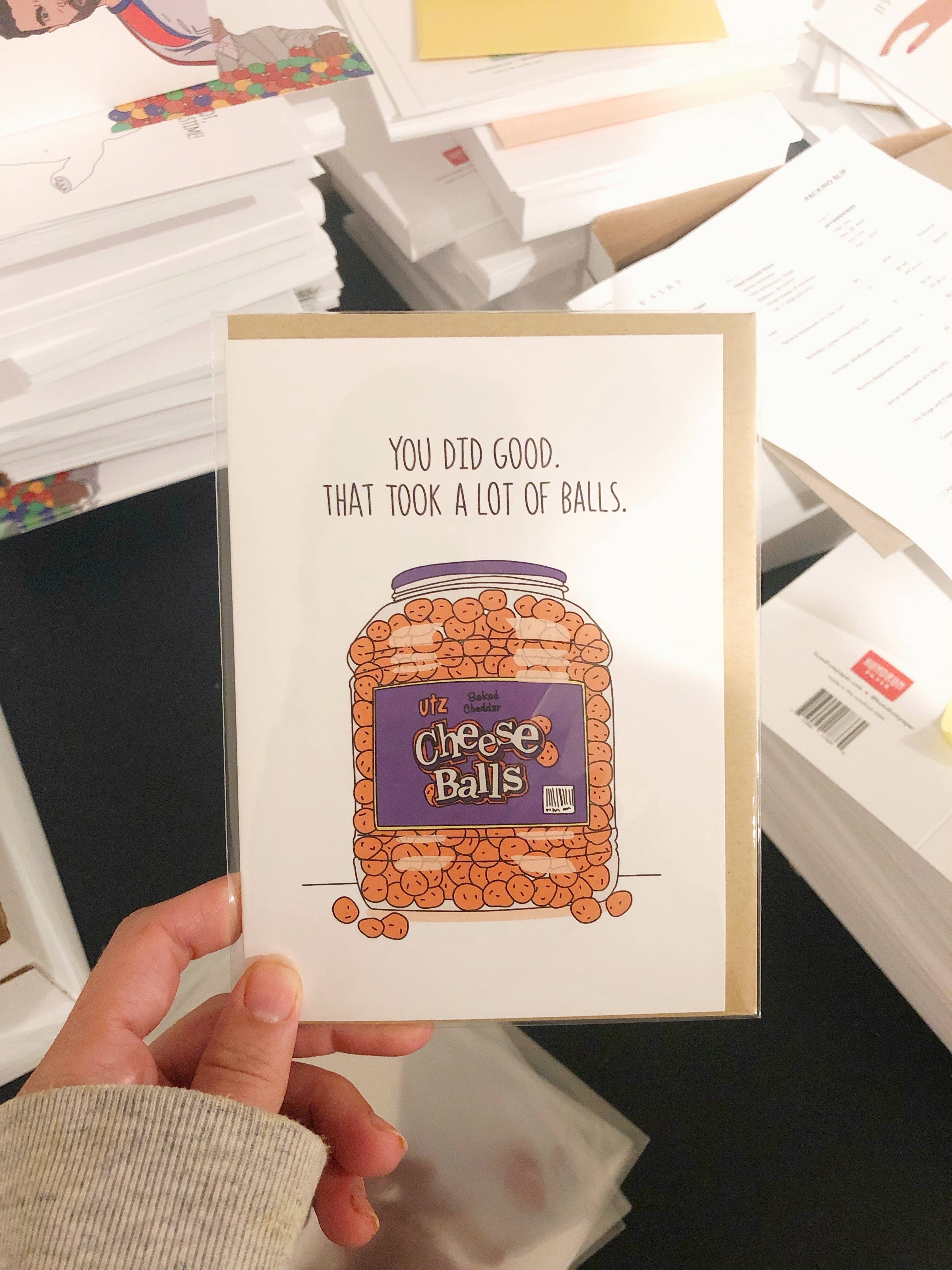 Card with a humorous design of a jar of cheese balls and text, held in front of a stack of papers.