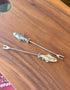 Two pewter fish picks with stainless steel prongs on a wooden board