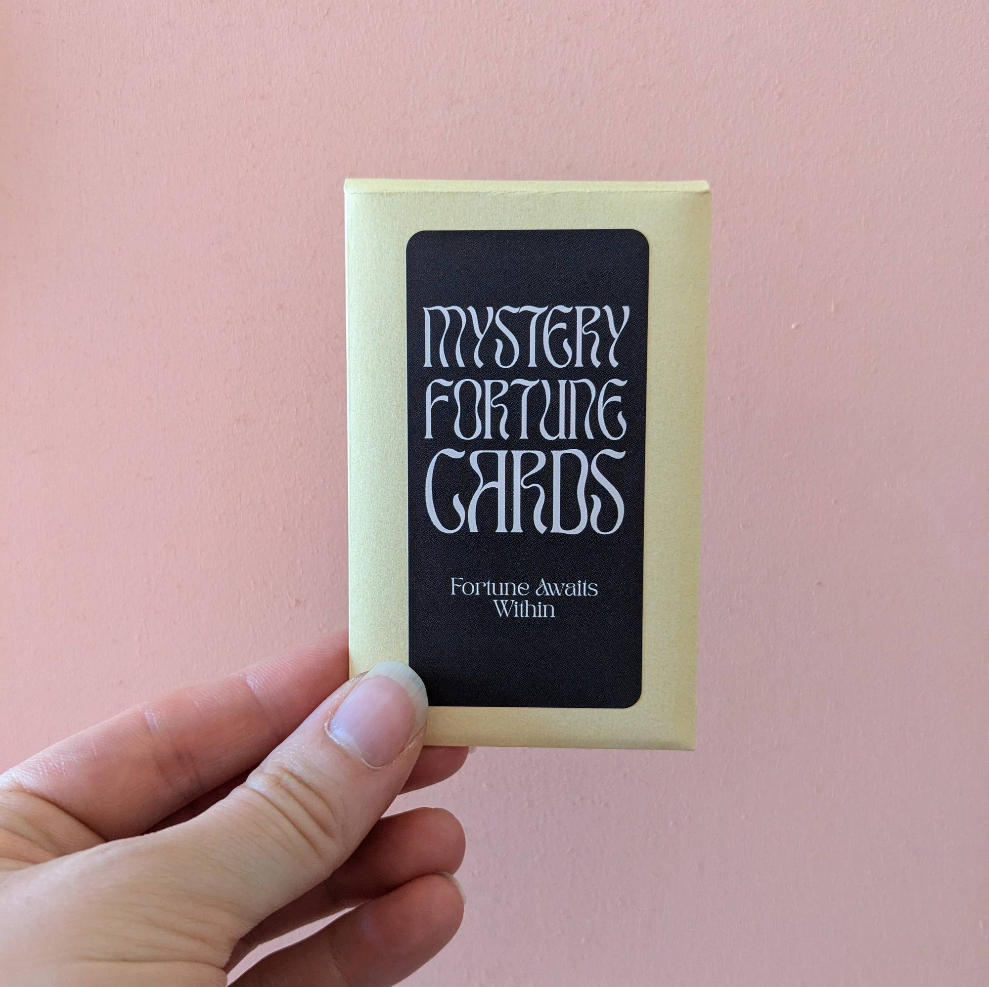 Hand holding a box of 'Mystery Fortune Cards' against a pink background