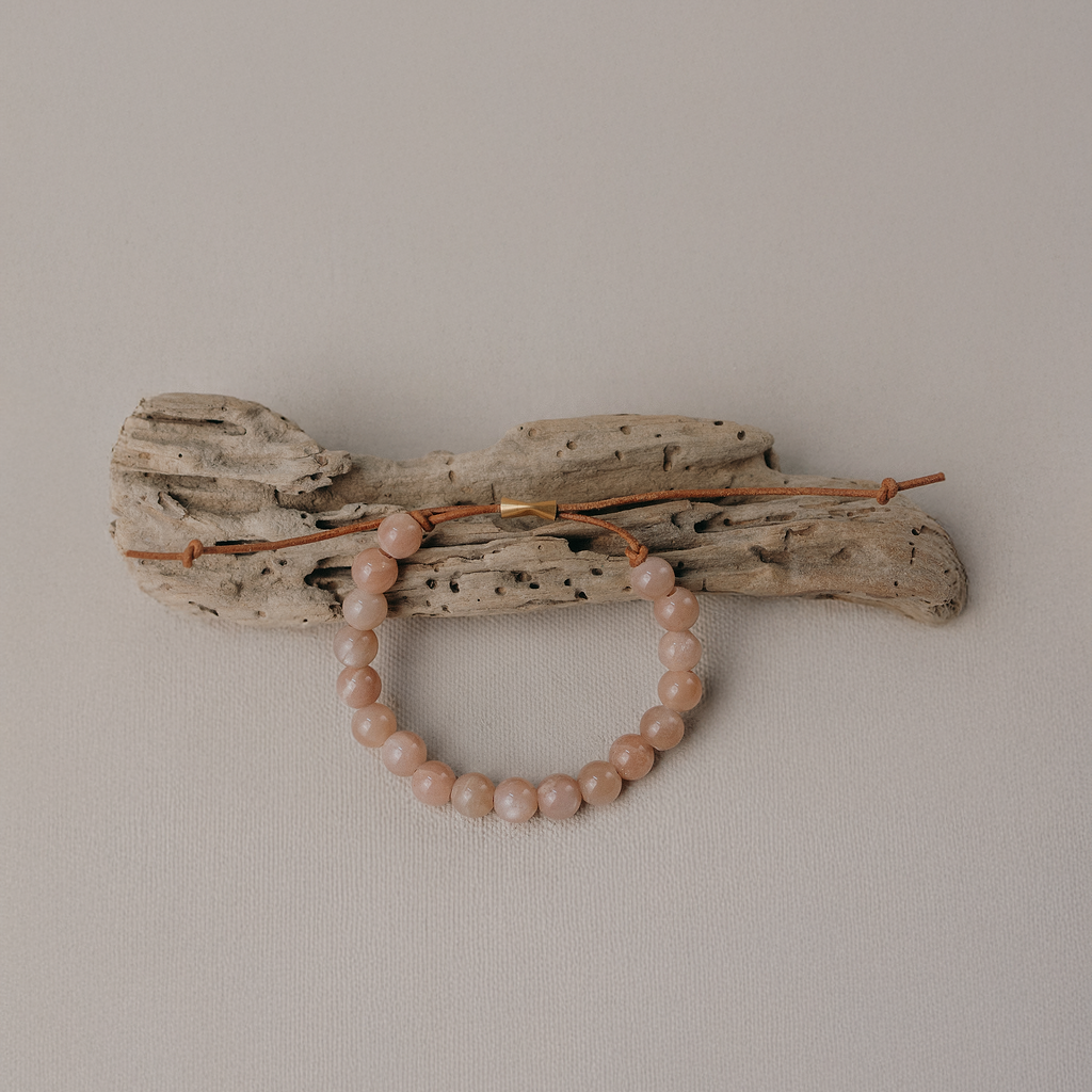 A beaded bracelet in soft blush pink rests on a piece of weathered driftwood with leather cord and 14K Gold-filled clasp set against a plain light gray background.