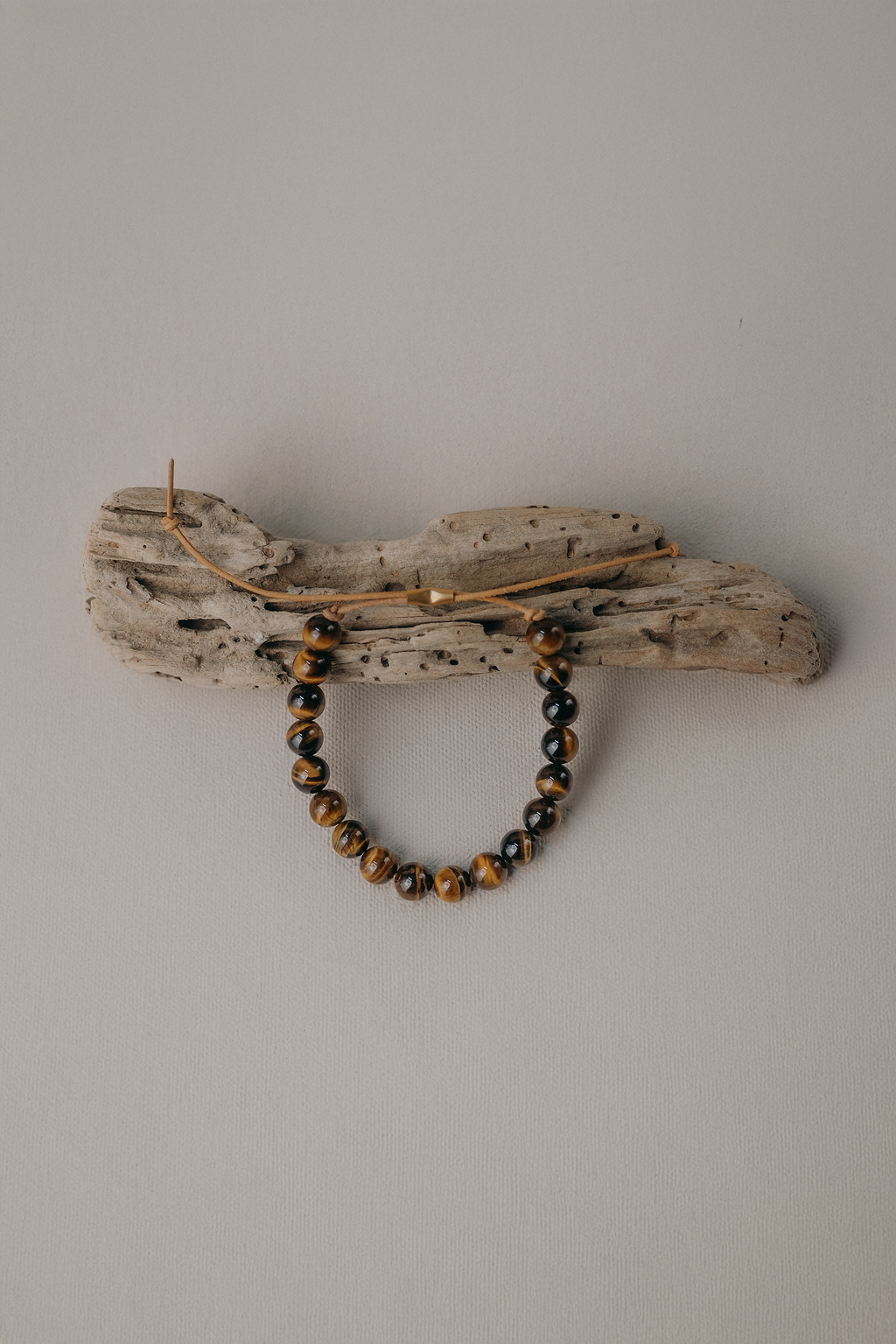 A beaded bracelet with brown and black marbled beads hangs from a rustic driftwood wall hook against a plain light gray wall.