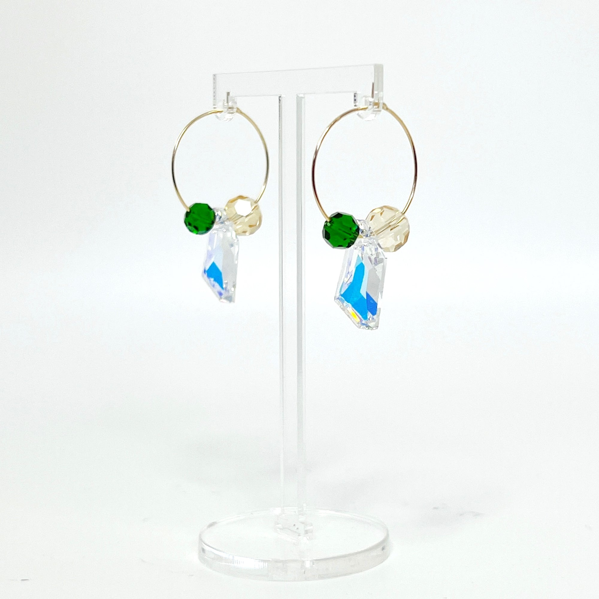 Understated elegance with a twist. The Small Muse Hoops combine a soft champagne crystal and a rich emerald green stone to frame an asymmetrical feature crystal at the center. The result is a beautifully balanced mix of refinement and individuality—perfect for the quietly powerful.