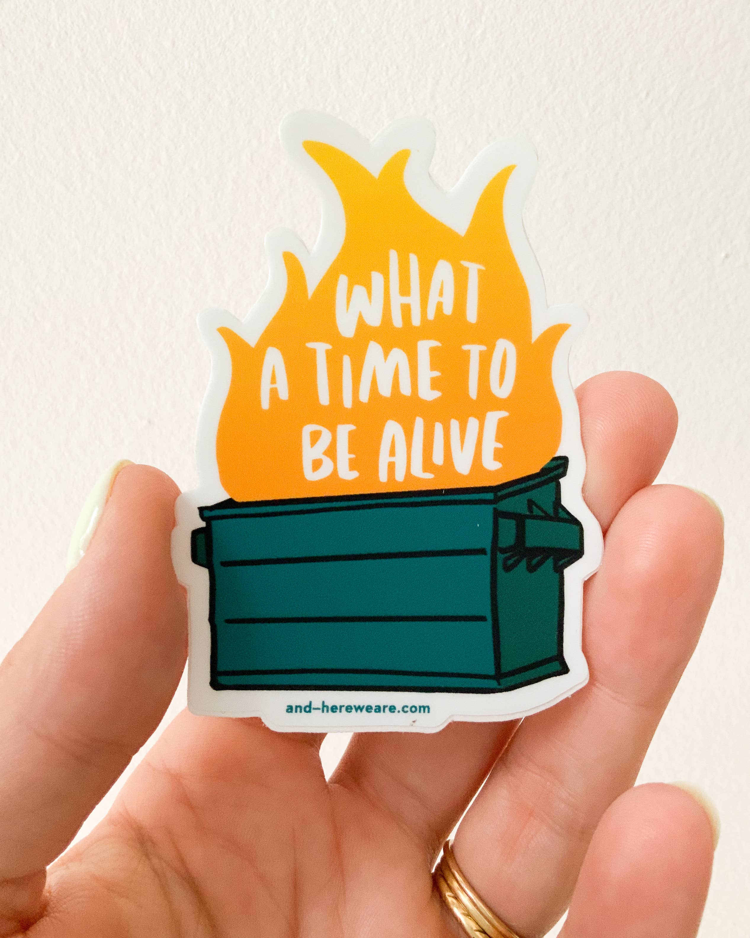 Sticker with a dumpster fire design and text held by a hand on a light background