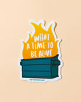 Sticker with a dumpster fire graphic and text 'What a time to be alive' on a beige background