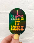 Sticker with 'HATE IT HERE' text held by a hand against a white background