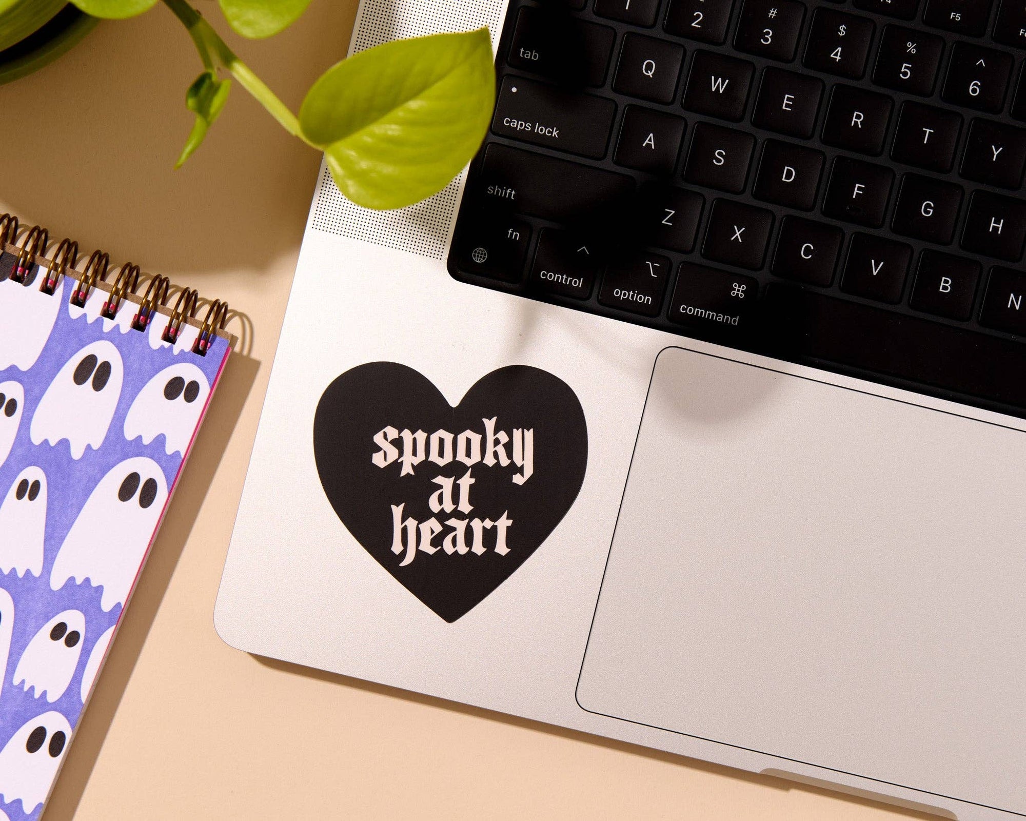 Laptop with a black heart-shaped sticker reading 'spooky at heart' on a desk with a notebook and keyboard.