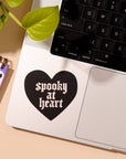Laptop with a black heart-shaped sticker reading 'spooky at heart' on a desk with a notebook and keyboard.