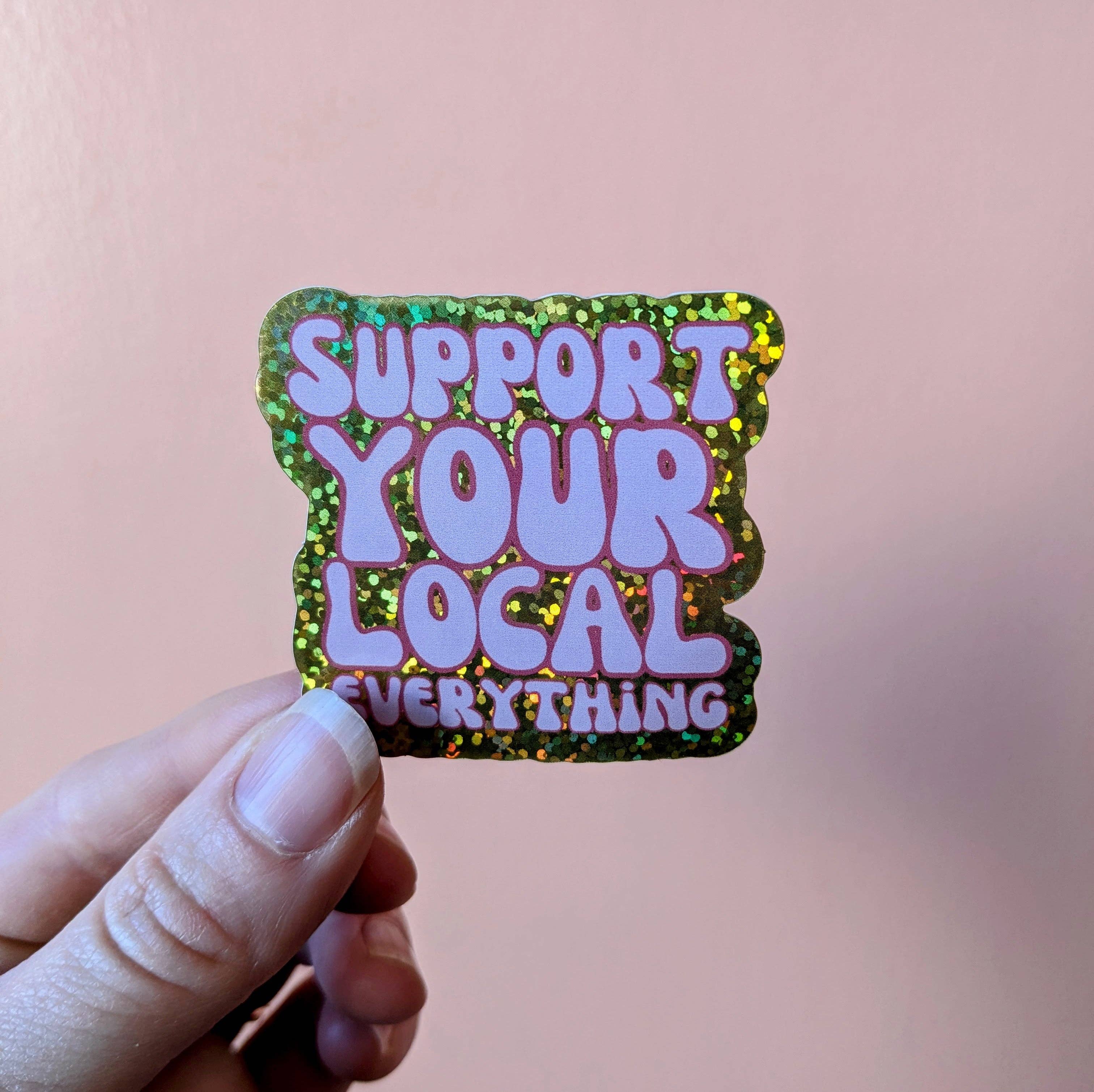 Holographic sticker with 'Support Your Local Everything' text held by a hand against a blurred background
