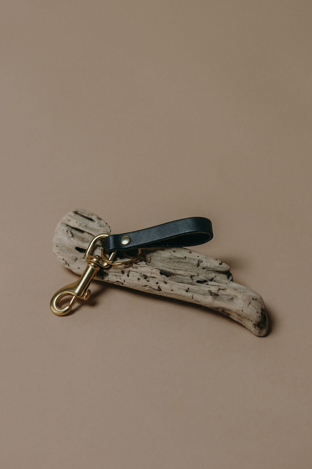 A rustic keychain or bag charm featuring a dark navy leather loop attached to a weathered, speckled off-white driftwood piece with a gold-tone metal swivel clasp, set against a solid warm beige background.