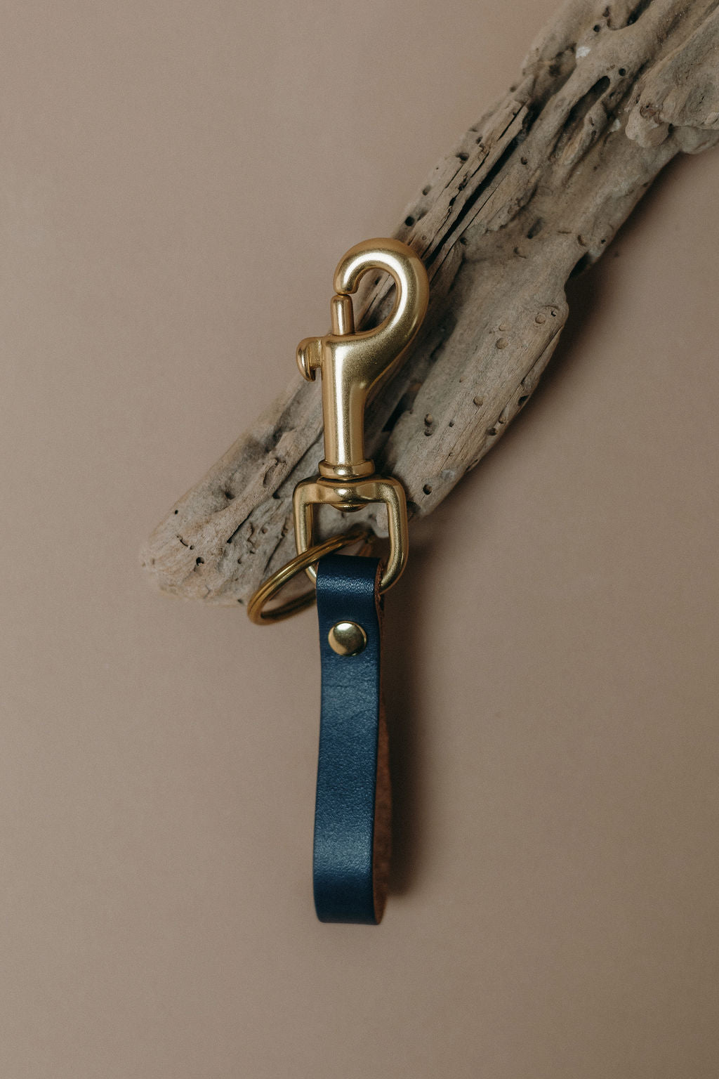 A dark blue leather key ring strap with a gold-tone metal swivel clasp and key ring, displayed against a neutral beige background with a piece of driftwood.