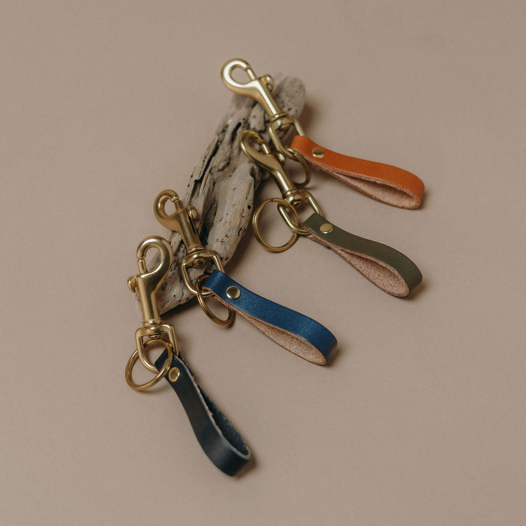 Four leather strap keychains in blue, orange, green, and gray, each attached to a gold-tone swivel clasp and displayed on a neutral beige background.