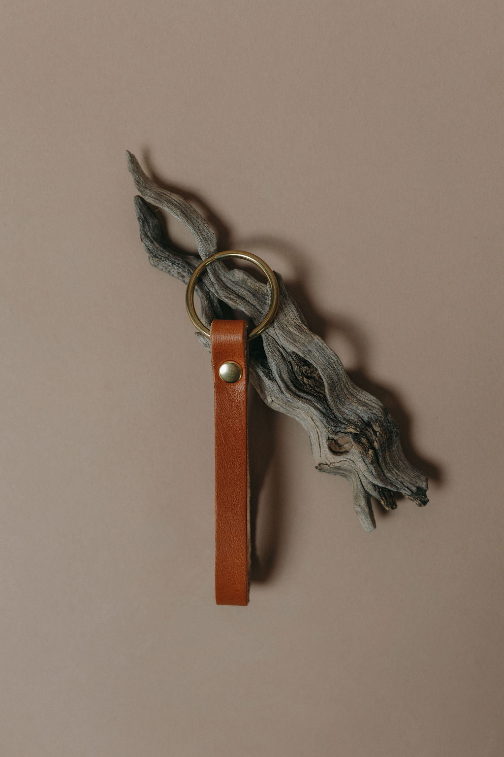 A brown leather key fob or keychain with a brass key ring, hanging from a piece of weathered driftwood against a solid taupe background.