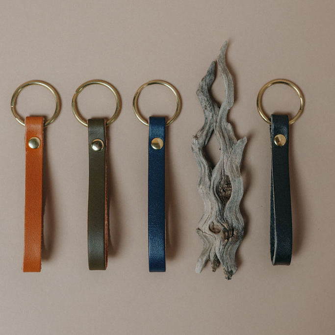 Four leather keychain loops in tan, olive, indigo and black, each attached to a round gold-tone key ring, arranged on a plain beige background with a decorative piece of driftwood between the blue and navy loops.
