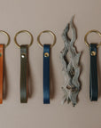 Four leather keychain loops in tan, olive, indigo and black, each attached to a round gold-tone key ring, arranged on a plain beige background with a decorative piece of driftwood between the blue and navy loops.