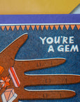 Greeting card with a hand holding gemstones and text 'You're a Gem' on a blue background.