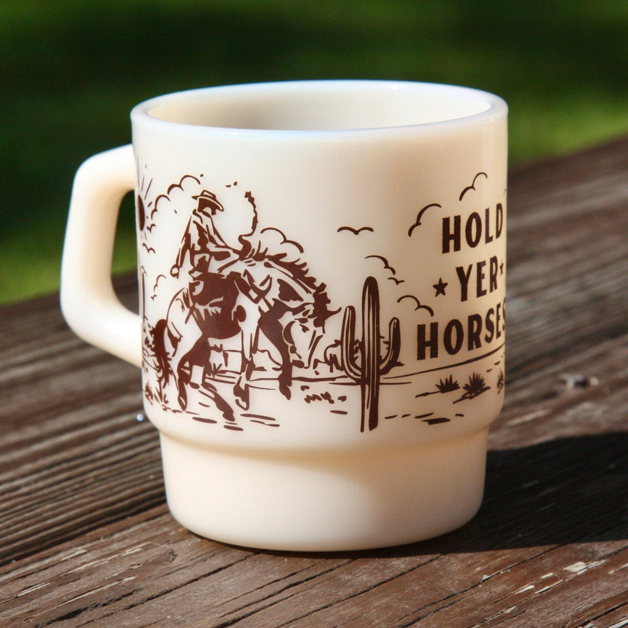 An off-white ceramic mug with a brown western-themed graphic and the text 'HOLD YER HORSES' sitting on a weathered wooden surface with a blurred green outdoor background.
